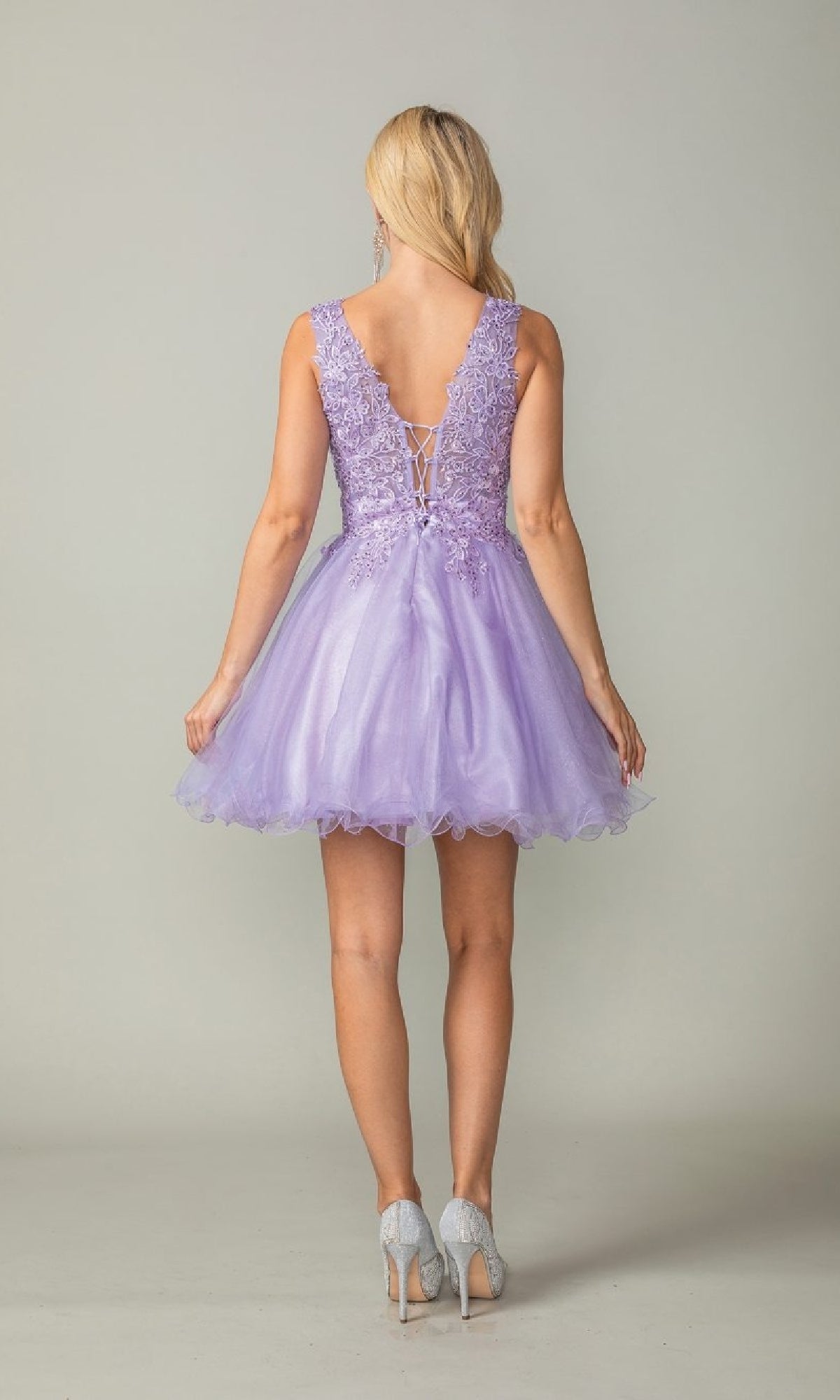 A woman faces away, wearing the Dancing Queen 3377 Tulle Babydoll Homecoming Dress in lavender with lace details, a V-back, and sleeveless design, paired with light gray heels.